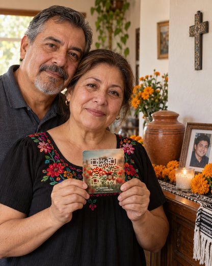 A man and a woman stand indoors together, holding a small medallion featuring a colorful QR code. The woman wears a black blouse with red and pink floral embroidery around the neckline.