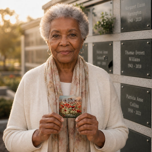 An elderly woman with short gray hair stands outdoors in front of memorial plaques, holding a small square QR medallion r with a colorful floral design