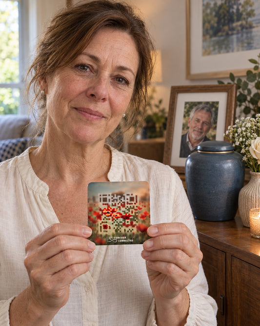 A woman in a white blouse holds up a small printed card featuring a colorful floral design and a QR code, with a home interior in the background including framed photos, a blue urn, and a candle.