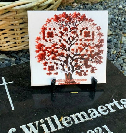 Tile with an QR code in the shape of a tree of life design on a stand, with a wicker basket and gravel in the background.